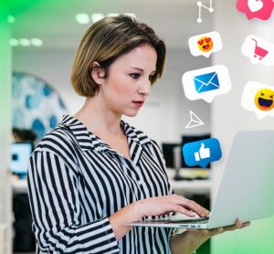 Mulher usando laptop em um ambiente de escritório, cercada por ícones de redes sociais representando curtidas, mensagens e carrinho de compras, simbolizando o trabalho de engajamento digital da Agência War para aumentar a interação e presença online.