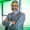 Homem de terno cinza e óculos, sorrindo com os braços cruzados em um ambiente corporativo, transmitindo confiança e profissionalismo.