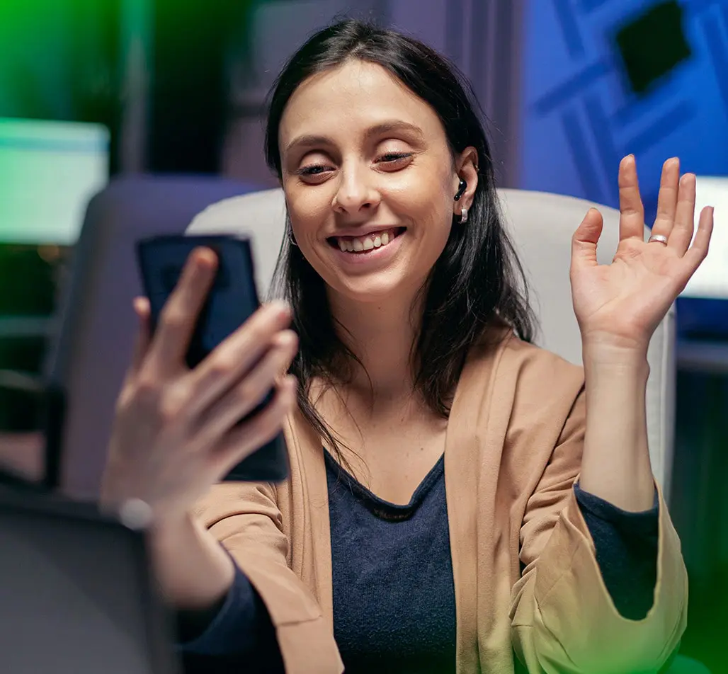 Mulher sorridente com fones de ouvido, participando de uma videochamada no celular. Ela acena com a mão enquanto olha para a tela, em um ambiente de escritório moderno, refletindo a abordagem acolhedora e profissional da Agência War em soluções de marketing digital.