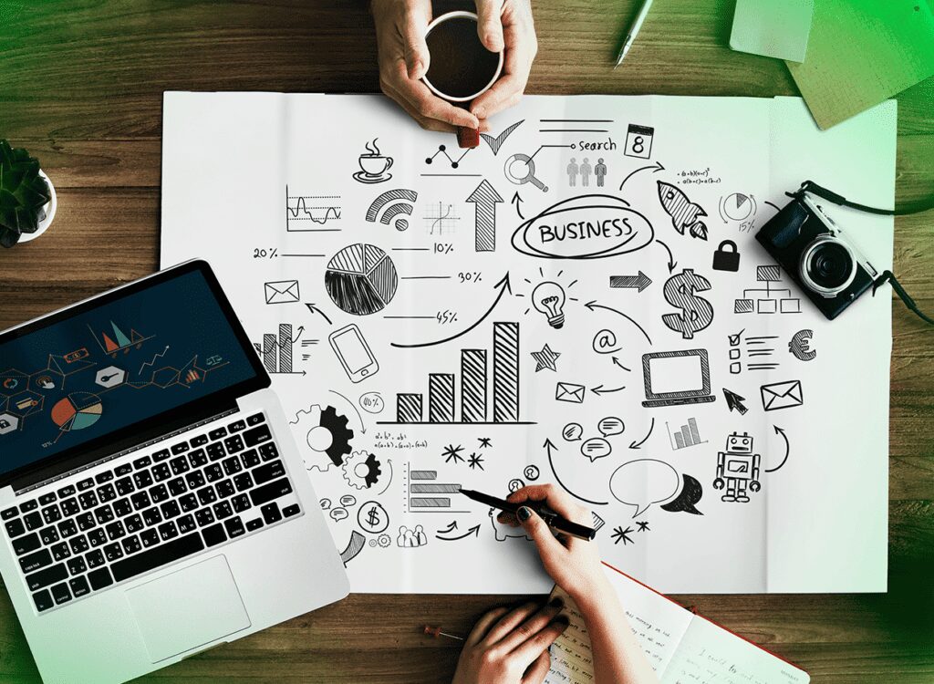 Mesa de trabalho com laptop, câmera, café e um grande papel com desenhos esquemáticos representando gráficos, crescimento de negócios, ícones de tecnologia e comunicação. Mãos seguram um café e uma caneta, destacando um brainstorming de planejamento estratégico.