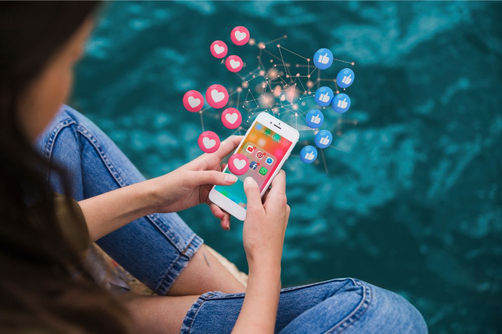 Pessoa sentada ao ar livre, segurando um smartphone que mostra aplicativos de redes sociais, enquanto ícones de curtidas e corações flutuam em volta, representando engajamento e interação online.