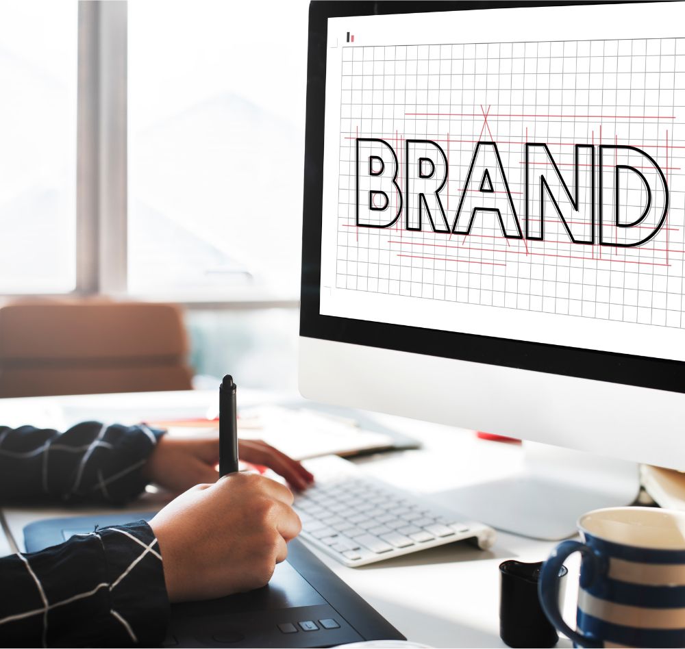 Pessoa usando uma caneta digital e uma mesa gráfica enquanto desenvolve a palavra 'BRAND' em uma tela de computador, sobre uma grade, representando o processo de criação de identidade visual.