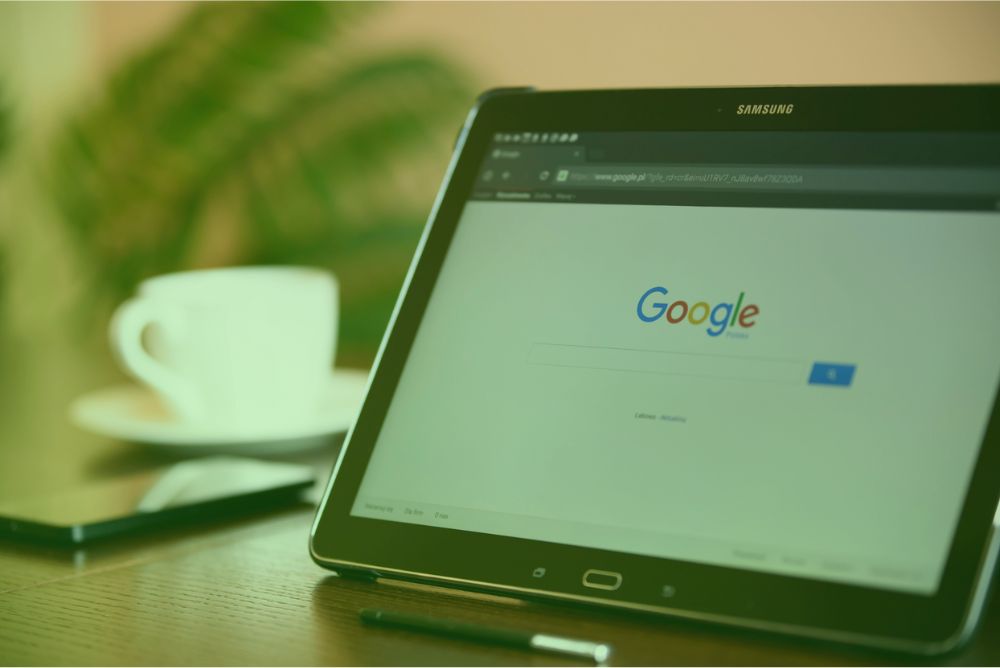 Tablet Samsung em cima de uma mesa, exibindo a página inicial de pesquisa do Google na tela. Ao fundo, há uma xícara branca e, ao lado, um smartphone e uma caneta.