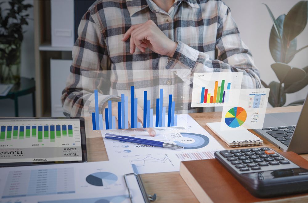 Pessoa sentada à mesa, vestindo camisa xadrez, analisando relatórios impressos e gráficos holográficos de barras e pizza, com calculadora e anotações ao redor, representando análise de dados e planejamento financeiro.