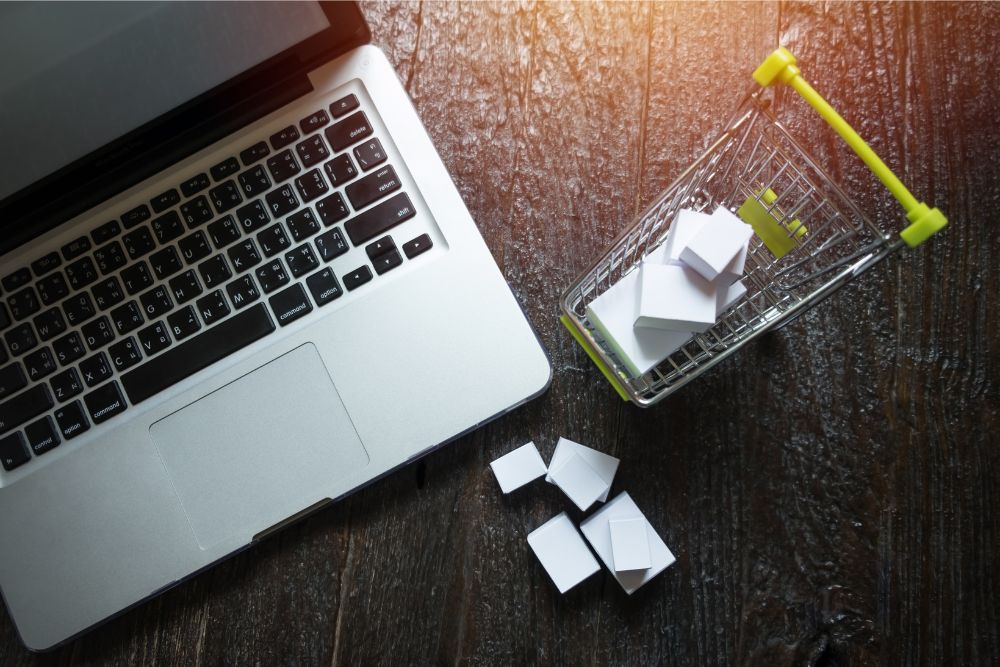 Laptop prateado sobre uma mesa de madeira, ao lado de um carrinho de compras em miniatura com pequenas caixas brancas, simbolizando compras online e comércio eletrônico.