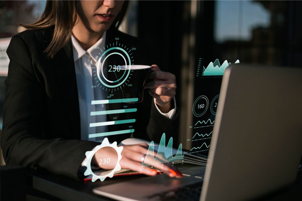 Mulher de terno trabalhando em um laptop enquanto visualiza gráficos e indicadores holográficos, tomando café ao mesmo tempo, simbolizando análise de dados e produtividade.