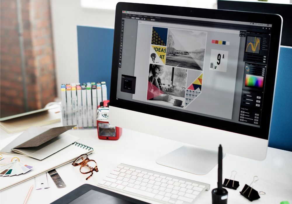 Computador de mesa exibindo um programa de design com colagens de imagens e blocos de cor, rodeado por materiais de escritório como cadernos, óculos, amostras de cor e uma caneta digital, representando o processo criativo de design gráfico.