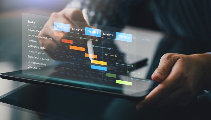 Pessoa segurando um tablet que exibe um gráfico de Gantt com tarefas listadas e barras coloridas ao longo de vários meses, representando planejamento e gerenciamento de projetos.